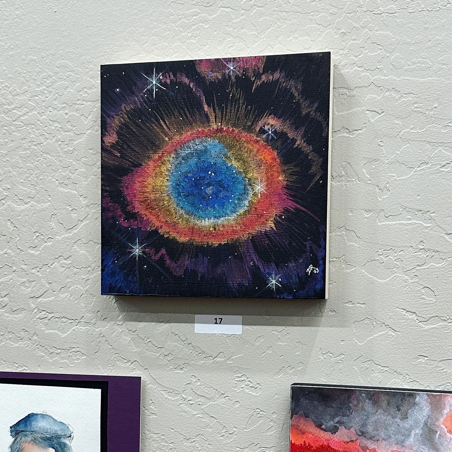 A black-primed square board with the Ring Nebula seen in shades of blue ringed with peach, gold, orange, red and pink, with scattered stars and eyelash-like rays in purple, bronze and pink, as seen by James Webb Space Telescope.