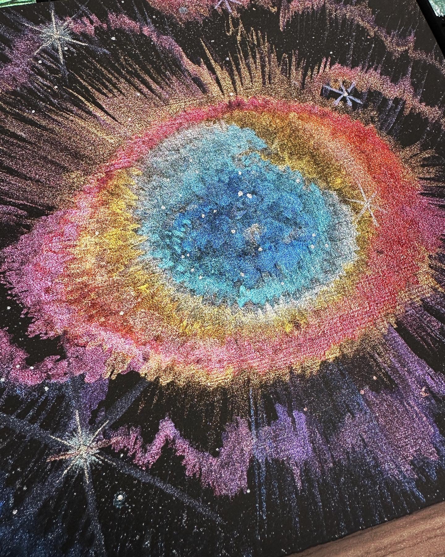 A black-primed square board with the Ring Nebula seen in shades of blue ringed with peach, gold, orange, red and pink, with scattered stars and eyelash-like rays in purple, bronze and pink, as seen by James Webb Space Telescope.